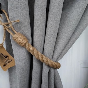 Pode incluir: Um amarrador de cortina de corda natural grossa com uma etiqueta castanha que diz "NEWSAILS". O amarrador est&aacute; a segurar cortinas cinzentas.