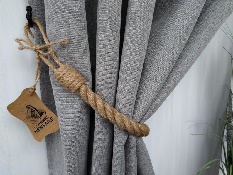 Pode incluir: Um amarrador de cortina de corda natural grossa com uma etiqueta castanha que diz "NEWSAILS". O amarrador est&aacute; a segurar cortinas cinzentas.