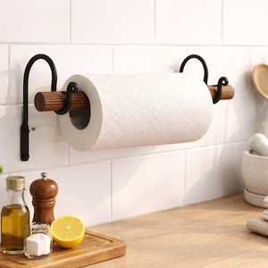 May include: A wall-mounted paper towel holder featuring a horizontal wooden rod supported by two black metal brackets with curved ends. The holder is mounted on a white tiled wall and holds a standard roll of white paper towels.