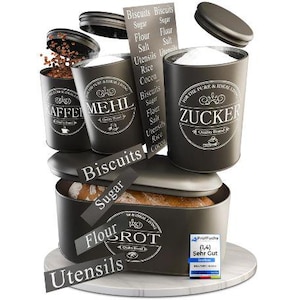 Peut inclure: Un ensemble de six boîtes en métal noir et blanc avec des étiquettes pour les biscuits, le sucre, la farine, le café et le sucre. Les boîtes sont empilées les unes sur les autres, avec une boîte à pain en bas. La boîte à pain a une étiquette qui dit "brot" et une étiquette qui dit "sehr gut".