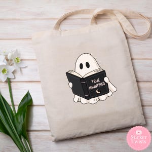 Może przedstawiać: Beżowa torba typu tote z kresk&oacute;wkowym duchem czytającym czarną książkę zatytułowaną "TRUE HAUNTINGS" z p&oacute;łksiężycem. Duch ma dwa czarne oczy i nosi białą prześcieradło. Torba ma długie, beżowe paski.