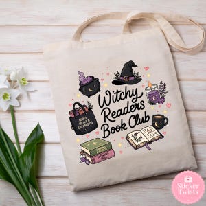 May include: Cream-colored tote bag featuring a whimsical design for a book club. The design includes a witch's hat, cauldron, books, a candle, and the text "Witchy Readers Book Club". The bag has a light beige strap.