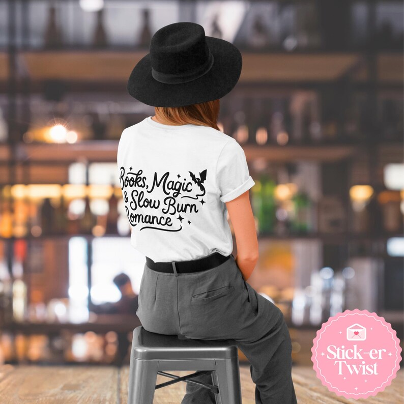 Puede incluir: Una camiseta blanca con el texto "Books, Magic & Slow Burn Romance" en escritura negra. La camiseta presenta un gr&aacute;fico negro de un hada y estrellas. La persona lleva un sombrero negro y pantalones grises.