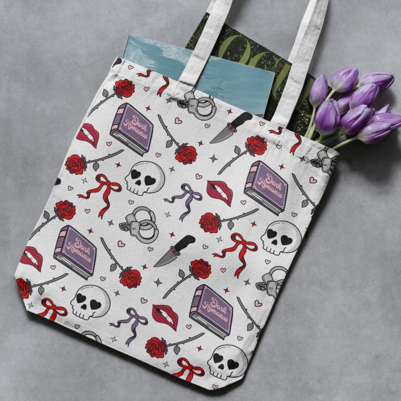 May include: White tote bag with a repeating pattern of skulls, roses, knives, lips, bows, and books with the text "Dark Romance". The bag has white handles and is filled with books and purple flowers.