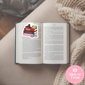 Puede incluir: Un libro abierto con un marcap&aacute;ginas que muestra una pila de libros con las etiquetas "Mafia Romance" y "Captured". El marcap&aacute;ginas tambi&eacute;n incluye una pistola, dinero y esposas. El libro est&aacute; sobre una superficie beige.