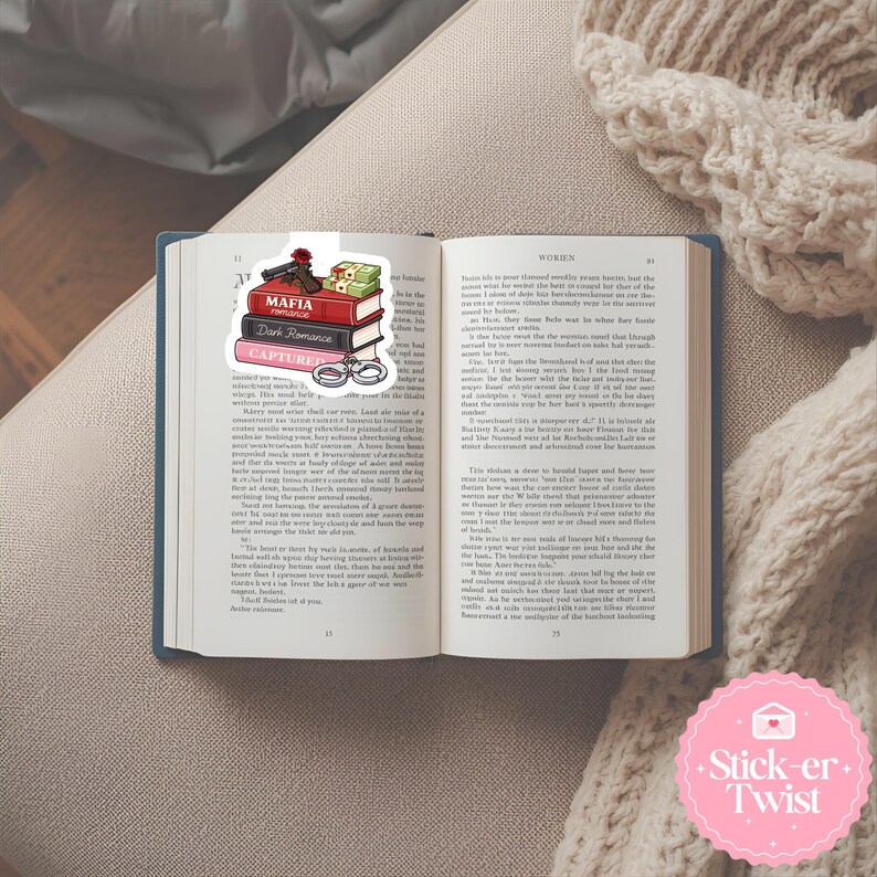 Puede incluir: Un libro abierto con un marcap&aacute;ginas que muestra una pila de libros con las etiquetas "Mafia Romance" y "Captured". El marcap&aacute;ginas tambi&eacute;n incluye una pistola, dinero y esposas. El libro est&aacute; sobre una superficie beige.