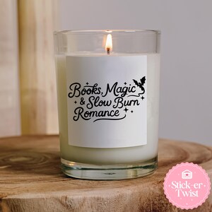 Puede incluir: Una vela encendida en un tarro de cristal transparente con una etiqueta blanca. La etiqueta dice "Books, Magic & Slow Burn Romance" en una fuente decorativa, con un gr&aacute;fico de murci&eacute;lago y estrellas. La vela est&aacute; sobre una superficie de madera.