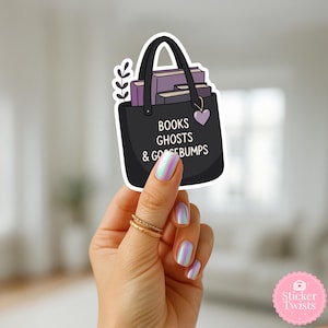 Może przedstawiać: Naklejka z czarną torbą typu tote z napisem "BOOKS GHOSTS & GOOSEBUMPS" i fioletowym sercem. Torba jest wypełniona fioletowymi książkami i gałązką liści. Naklejka jest trzymana przez dłoń ze złotymi pierścionkami i opalizującymi paznokciami.