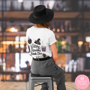 May include: White t-shirt with the text "Witchy Readers Book Club" and witch-themed illustrations. The design includes a cauldron, witch hat, and other magical elements. The person is wearing a black hat and gray pants.