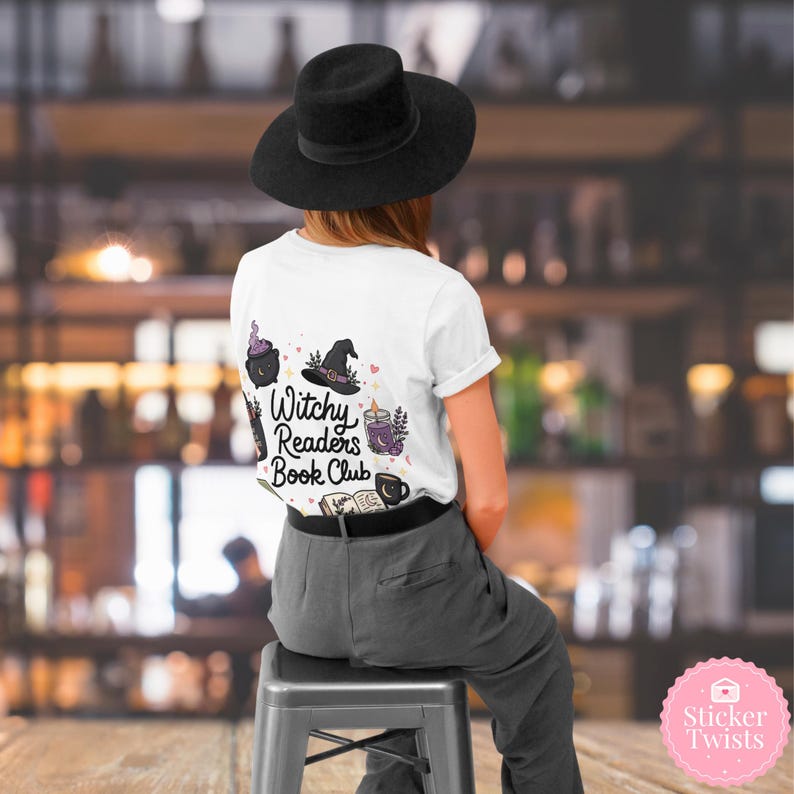 May include: White t-shirt with the text "Witchy Readers Book Club" and witch-themed illustrations. The design includes a cauldron, witch hat, and other magical elements. The person is wearing a black hat and gray pants.