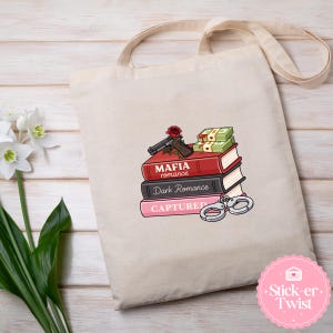 Puede incluir: Bolso tote beige con un dise&ntilde;o que presenta una pila de libros con los t&iacute;tulos "Mafia Romance", "Dark Romance" y "Captured". Una pistola, una rosa, dinero y esposas est&aacute;n encima. El bolso tiene correas largas y un dise&ntilde;o gr&aacute;fico.