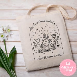 May include: Beige canvas tote bag with a black line art design. The design features a castle, sword, roses, and the text "her heart was made for dark romance". The bag has long, beige straps.