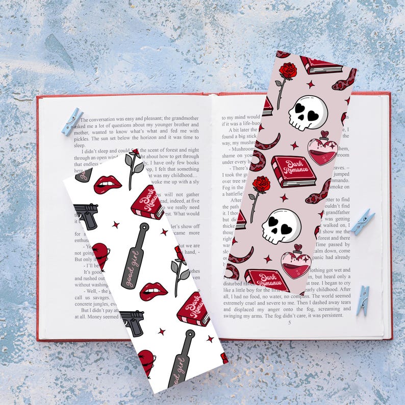 May include: Two bookmarks with a book, skull, rose, and heart design. One bookmark has a good girl design, and the other has a dark romance design. The bookmarks are on top of an open book.