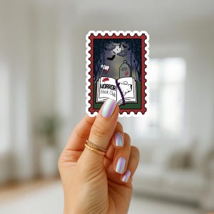 May include: A sticker in the shape of a postage stamp with a horror theme. The design includes a book with the words "HORROR Book Club", a ghost, an axe, a tombstone, and a full moon. The sticker is held by a hand with iridescent nails and gold rings.