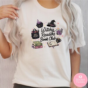 May include: White t-shirt featuring a colorful graphic design for a book club. The design includes a witch's hat, cauldron, books, a candle, and the text "Witchy Readers Book Club". The design is surrounded by small hearts.