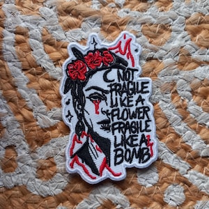 Peut inclure: Écusson brodé blanc avec un profil de femme, une couronne florale et des détails rouges. Le patch porte l'inscription "NOT FRAGILE LIKE A FLOWER FRAGILE LIKE A BOMB" en noir et rouge.