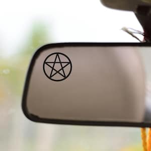 May include: A black rearview mirror with a pentagram sticker on the glass. A gold and orange tassel hangs below the mirror. The background is blurred, showing the interior of a vehicle.