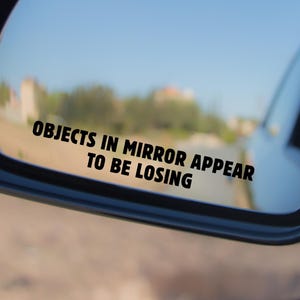 May include: Black side mirror of a vehicle with the text "OBJECTS IN MIRROR APPEAR TO BE LOSING" in black. The mirror reflects a blurred outdoor scene with a blue sky.