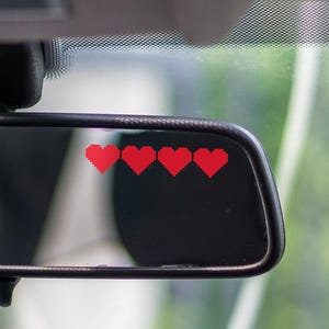 May include: A car's rearview mirror with four red heart stickers on the right side. The mirror is rectangular with rounded corners and a black frame. The background is blurred, showing green foliage.