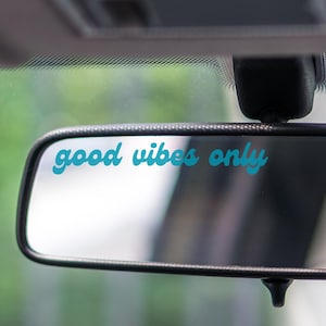 May include: A black rearview mirror with the words "good vibes only" in teal script. The mirror is mounted inside a vehicle, with a blurred green background visible through the windshield.