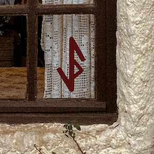 May include: A rustic window with a dark brown frame and a white lace curtain. A red symbol is painted on the curtain. The window is set in a textured, off-white wall, with a dark brown wooden shutter on the right.