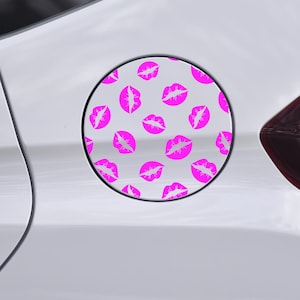May include: A white car with a circular gas cap cover featuring a pattern of bright pink lips on a light gray background. The car's door handle and taillight are visible.