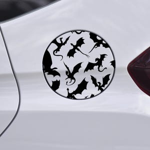 May include: A circular black and white decal featuring multiple silhouettes of dragons in various poses. The decal is affixed to the side of a white vehicle, near the fuel door and a red tail light.