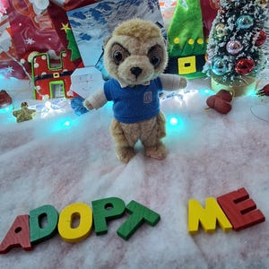 Puede incluir: Un peluche de suricato con un suéter azul, de pie sobre una superficie nevada. Las palabras "ADOPT ME" están escritas con letras de madera de colores. Decoraciones navideñas, incluido un pequeño árbol, están en el fondo.
