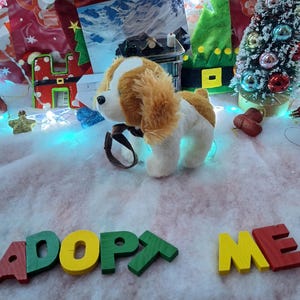 Puede incluir: Un perro de peluche con pelaje marrón y blanco, con un collar y correa marrones. Las palabras "ADOPT ME" están escritas con letras de madera de colores. La escena está ambientada sobre una superficie nevada con adornos navideños.