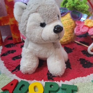 Puede incluir: Un peluche de perro husky gris y blanco con ojos y nariz negros. El juguete está sentado sobre una alfombra roja con un diseño de sandía. La palabra "ADOPT" está escrita con letras de madera de colores.