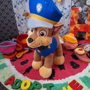 Può includere: Un peluche di un cane dei cartoni animati con cappello e uniforme blu della polizia. Il cane è marrone e marrone chiaro, con un naso nero e grandi occhi. Il giocattolo è su un tappeto rosso e verde con le parole "ADOPT ME" in lettere di legno colorate.