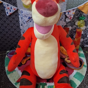 May include: A large Tigger plush toy from Winnie the Pooh. The soft toy is mainly orange with black stripes, a cream-coloured belly, and a pink mouth. It has large, round eyes and a red nose. The toy is sitting on a colourful rug.