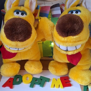 Puede incluir: Dos juguetes de peluche de perro amarillos con narices marrones y lenguas rojas, sonriendo y levantando sus patas. Los juguetes están sentados sobre una superficie blanca con la palabra "ADOPT ME" escrita en letras de madera de colores.