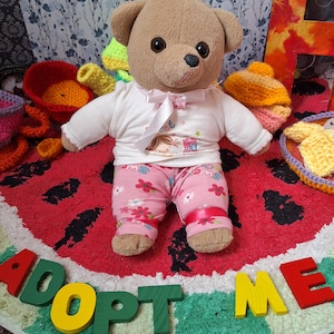 Puede incluir: Un oso de peluche marrón vestido con una camiseta blanca con un lazo rosa y pantalones florales. El oso está sobre una alfombra roja con forma de sandía con las palabras "ADOPT ME" en letras de madera de colores.