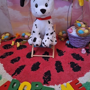 Puede incluir: Un perro dálmata de peluche con manchas negras y un collar rojo está sentado en una pequeña silla de madera. El perro está sobre una alfombra roja con forma de sandía con las palabras "ADOPT ME" en letras de madera coloridas.