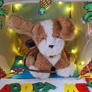 May include: A brown and white plush dog toy with a brown leather collar sits inside a light brown canvas tote bag. The bag is decorated with a pineapple, palm trees, and string lights. The words "ADOPT ME" are spelled out in colorful wooden letters in front of the bag.