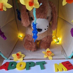 May include: A brown and white plush dog toy sits inside a cardboard house decorated with yellow flowers and string lights. The house has a sign that reads "Adopt Me".