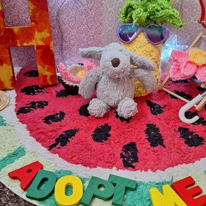Puede incluir: Un juguete de peluche gris con forma de perro está sentado sobre una alfombra con forma de sandía con semillas negras. También se ve una piña de ganchillo con gafas de sol en forma de corazón y las palabras "ADOPT ME" en letras de madera de colores.