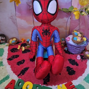 Puede incluir: Muñeco de peluche de Spider-Man con un disfraz rojo y azul con un emblema de araña negra. El muñeco está sentado en una alfombra con temática de sandía con las palabras "ADOPT ME" en letras coloridas. Decoraciones de Pascua en el fondo.