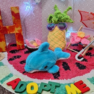 Puede incluir: Un delfín azul de peluche descansa sobre una alfombra con diseño de sandía, rodeado de otros artículos hechos a mano. Una piña de ganchillo con gafas de sol, una silla de playa en miniatura y las palabras "ADOPT ME" en letras de madera coloridas.