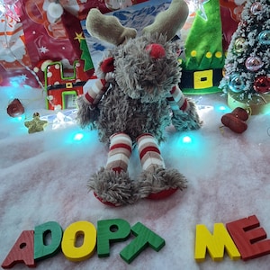 Puede incluir: Un peluche de reno con una nariz roja, bufanda y piernas a rayas. Las palabras "ADOPT ME" están escritas con letras de madera de colores frente al reno. Decoraciones y luces navideñas en el fondo.