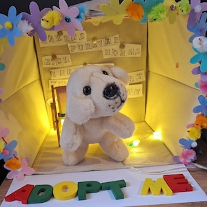 Puede incluir: Un juguete de peluche de perro blanco dentro de una caja de cartón decorada con flores de colores y las palabras "Adoptame". La caja también tiene las palabras "Forever Homes" y "Etsy" escritas en ella.