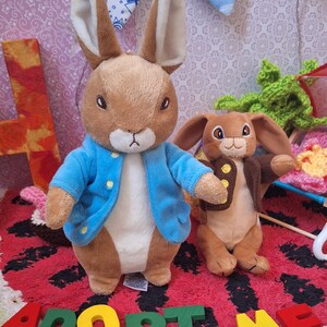 Puede incluir: Dos conejos de peluche. Uno es marrón y beige, con una chaqueta azul con botones dorados. El otro es más pequeño, marrón y lleva una chaqueta marrón. Las palabras "ADOPT ME" están escritas con letras de madera de colores.