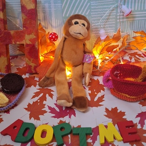 Puede incluir: Un mono de peluche marrón con una etiqueta, sentado entre hojas de otoño y luces de hadas. Las palabras "ADOPT ME" están escritas con letras de madera de colores. También se ve una pequeña cesta de ganchillo y una "H" decorativa.