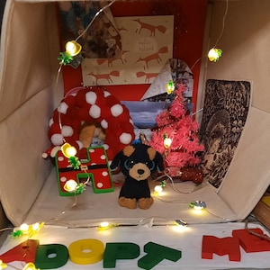 Puede incluir: Una caja blanca con una letra H de lunares rojos, blancos y azules, un árbol de Navidad rosa, un perro de peluche negro y blanco, una corona roja y blanca y una cadena de luces que deletrean "ADOPT ME".
