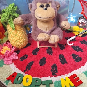 Puede incluir: Un peluche de mono con pelaje marrón y cara beige sentado en una pequeña silla de madera. La escena incluye una piña de ganchillo, una flor rosa, un mango de paraguas blanco y una alfombra redonda con patrón de sandía y el texto ADOPT ME.