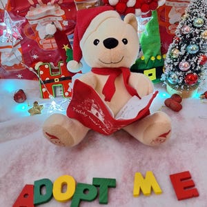 Op de afbeelding: Een beige teddybeer met een kerstmuts en rode sjaal leest een rood boek. De scène omvat een kleine kerstboom en de woorden "ADOPT ME" in kleurrijke houten letters.