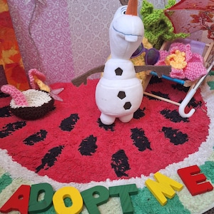 Puede incluir: Un peluche de Olaf con gafas de sol, una silla de playa en miniatura y una flor de ganchillo sobre una alfombra con temática de sandía. La palabra "ADOPT ME" está escrita con letras de madera de colores.