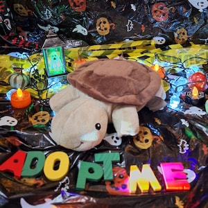 Puede incluir: Un peluche de tortuga con caparazón marrón y cuerpo beige claro descansa sobre un fondo de Halloween. La escena incluye calabazas decorativas, una linterna brillante con un fantasma y las palabras "ADOPT ME" en letras coloridas.