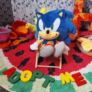 Puede incluir: Un peluche de Sonic the Hedgehog está sentado en una silla de playa en miniatura sobre una alfombra con temática de sandía. La alfombra tiene un fondo rojo con formas de semillas negras. Las palabras "ADOPT ME" están escritas en letras de madera de colores.